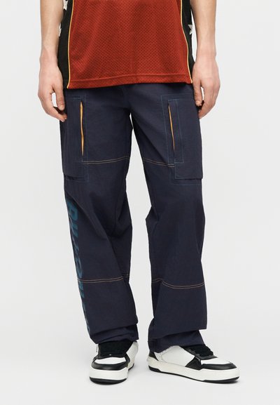 Άτομο που φορά χαλαρό σκούρο cargo παντελόνι με πορτοκαλί ανοίγματα στις τσέπες, κόκκινο τοπ από δίχτυ και μαύρα και λευκά αθλητικά παπούτσια.