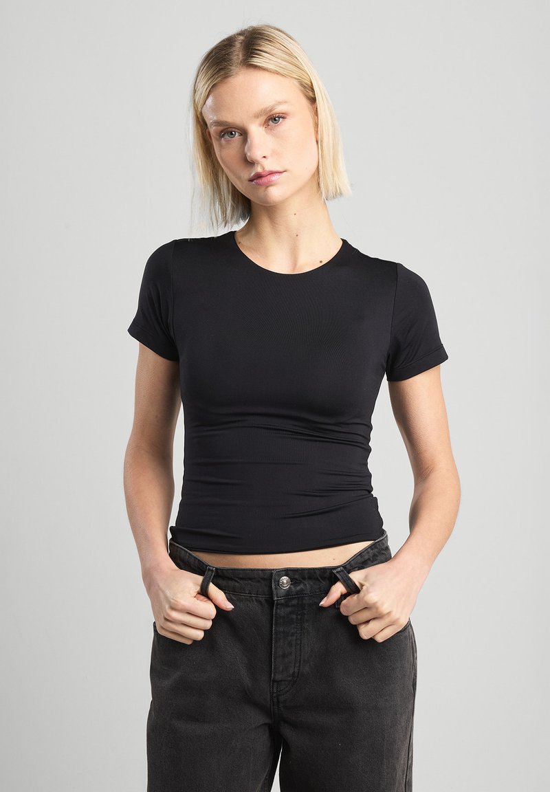 Black fitted T-shirt made of ribbed fabric; short sleeves, crew neck, and gathered detailing at the waist. Worn with black high-waisted jeans.