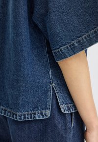 Détail de la manche et du côté d'une chemise en denim bleu avec une petite fente latérale portée sur un pantalon en denim assorti, montrant une partie d'un bras nu.