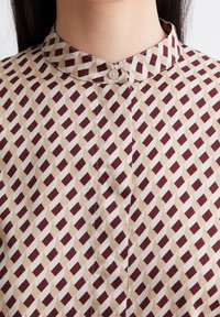 Bluzka z kołnierzykiem w geometryczny wzór w kolorze burgundowym i kremowym. Wykonana z lekkiego materiału o miękkiej fakturze, z zapięciem na guziki.