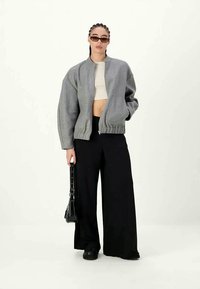 Gray, cropped bomber jacket with a high collar, ribbed hem, and wide black pants; model holding a black bag, wearing sunglasses.