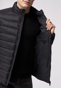Giacca a vento nera con texture trapuntata, colletto alto e tasche con zip. Aperta per rivelare una fodera interna liscia. Top scuro sotto.