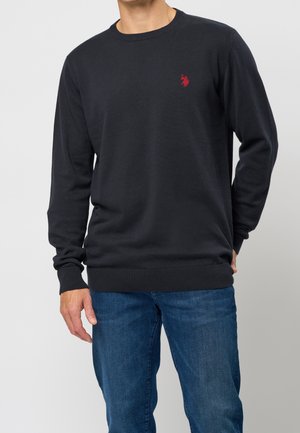 Homme portant un pull noir à manches longues et col rond avec un petit logo rouge sur la poitrine et un jean bleu, debout devant un fond uni.