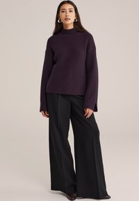 Femme portant un pull en maille ribbed violet foncé avec des manches fendues et un pantalon noir à jambes larges, se tenant contre un fond beige uni.