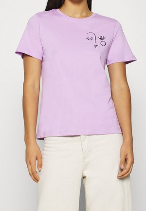 Frau trägt ein fliederfarbenes T-Shirt mit kurzen Ärmeln und einem schwarzen abstrakten Gesichtsmotiv auf der linken Brust, kombiniert mit weißer Hose.