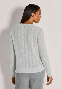 Frau mit langen dunklen Haaren, die einen hellgrauen strukturierten Strickpullover und graue Hosen trägt, von hinten vor einem schlichten Hintergrund gezeigt.