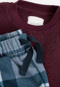 Tummanpunainen fleece-huppari, jossa on tekstuuri pinta, yhdistettynä sinivalko-harmaisiin ruudullisiin pehmeisiin kudottuihin housuihin, joissa on tummat nyörit.