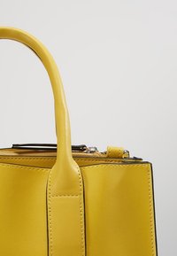 Sac à main en cuir jaune avec une anse arrondie unique et une fermeture éclair visible sur le dessus, sur un fond clair uni.