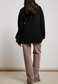 Schwarzer gerippter Oversize-Pullover mit langen Ärmeln, kombiniert mit hellbraunen weit geschnittenen Hosen. Fokus auf die Textur und die bequeme Passform der Kleidungsstücke.