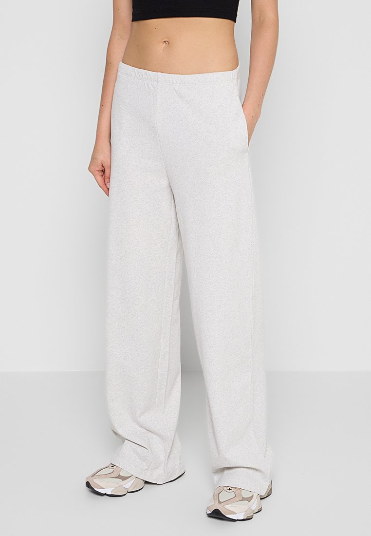 Donna che indossa pantaloni sportivi larghi grigi chiari, un crop top nero e sneakers beige bianche, in piedi contro uno sfondo neutro.