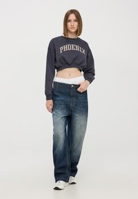 Czarno-szary skrócony sweatshirt z napisem "PHOENIX", połączony z luźnymi niebieskimi dżinsami z stopniowym wyblaknięciem, noszonymi ponad talią.