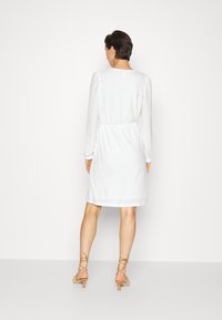 Vestido branco com mangas longas, tecido texturizado, cintura franzida, comprimento até ao joelho, combinado com sandálias bejes com tiras.