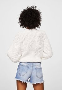 Suéter blanco de punto con cuello alto y mangas anchas, combinado con shorts vaqueros azules con dobladillos deshilachados y bolsillos traseros.