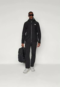 Jachetă waterproof neagră cu fermoar frontal și capușă ajustabilă, purtată împreună cu pantaloni negri asortați și pantofi gri. Rucsac negru inclus.