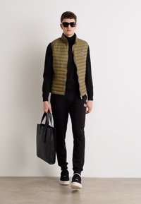 Colete acolchoado verde-oliva sobre um suéter de gola alta preto, combinado com calças de moletom pretas, sapatos pretos e uma bolsa tote preta. Texturas suaves e design em camadas.