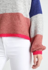 Farbblockpullover in Grau, Pink, Blau und Weiß. Hergestellt aus weichem, flauschigem Material mit gerippten pinken Bündchen und Saum. Weiße Jeans sichtbar.