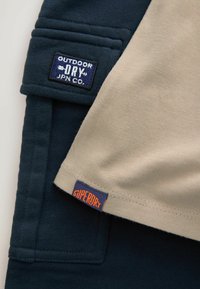 Námořnická modrá a béžová látka s dvěma štítky na oblečení: "Outdoor Dry JPN Co." modře na námořnické a "Superdry" oranžově na béžové.