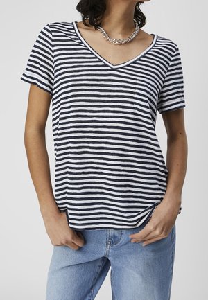 Kobieta ubrana w czarno-biały, poziomo w paski t-shirt z dekoltem w serek i krótkimi rękawami oraz jasnoniebieskie dżinsy z srebrnym naszyjnikiem z łańcuszka.