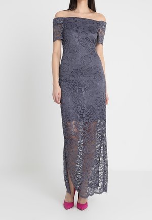 Kvinde iført en grå off-shoulder blondekjole med blomsterprint og gennemsigtig nederdel, kombineret med lyserøde høje hæle.