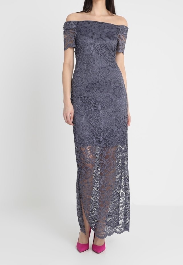 Femme portant une robe en dentelle grise à épaules dénudées avec motifs floraux et une jupe inférieure transparente, associée à des talons hauts rose vif.