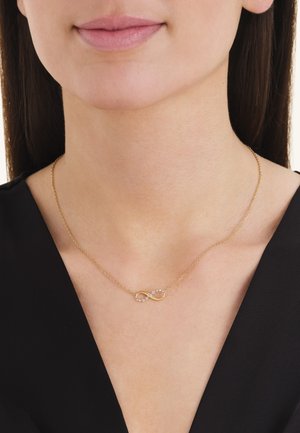 Collar de infinito de oro con pequeños diamantes, llevado en el cuello de una mujer, combinado con un top negro de escote en V.