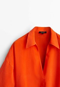 Col et partie supérieure d'une chemise orange vif à col avec une étiquette de marque noire visible à l'intérieur de l'encolure.