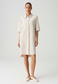 Beiges, knopfbares Hemdkleid mit kurzen Ärmeln, zwei Brusttaschen und lockerer Passform. Geschmeidige Textur und mittellanger Saum, geeignet für den Freizeitlook.