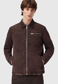 Brązowa kurtka z płótna z zamkiem z przodu, dwoma bocznymi kieszeniami i kieszenią na piersi. Posiada kołnierz z sztruksu i postrzępione detale u dołu.