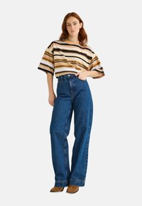 Chemise à manches courtes rayée beige, marron et blanc ; associée à un jean en denim bleu taille haute ; le mannequin pose avec une main sur la hanche.