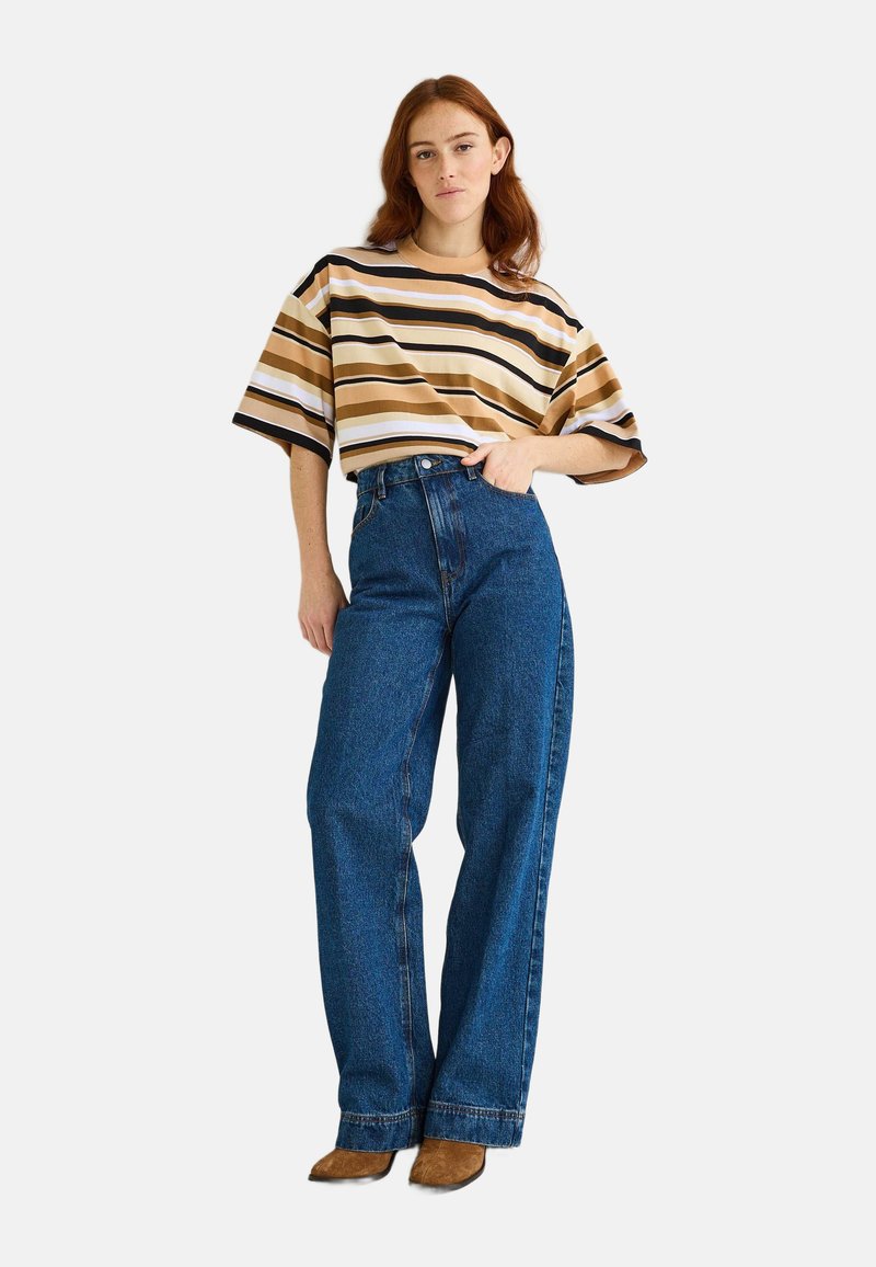 Chemise à manches courtes rayée beige, marron et blanc ; associée à un jean en denim bleu taille haute ; le mannequin pose avec une main sur la hanche.