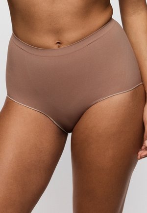 Vrouw die naadloos lichtbruin ondergoed met hoge taille draagt en ontspannen met één arm langs haar lichaam staat tegen een effen achtergrond.