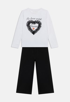 Biały top z długim rękawem z czarnym falbaniastym sercem i napisem "Zakochana w Guess" w połączeniu z szerokimi czarnymi spodniami.
