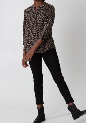 Person iført en sort blomstret langærmet bluse, sorte smalle bukser og sorte ankelstøvler, stående mod en ensfarvet lys baggrund.