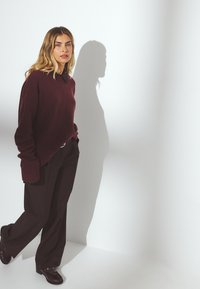 Burgundijas krāsas adīts džemperis ar pārspīlētu piegriezumu un ribotām piedurknēm, kombinēts ar tumšiem platām kājām biksēm un spīdīgiem mokasīniem, radot maigu ēnu.