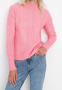 Pull en tricot rose avec un col haut, présentant un motif texturé. Les manches sont longues et il est associé à un jean bleu.