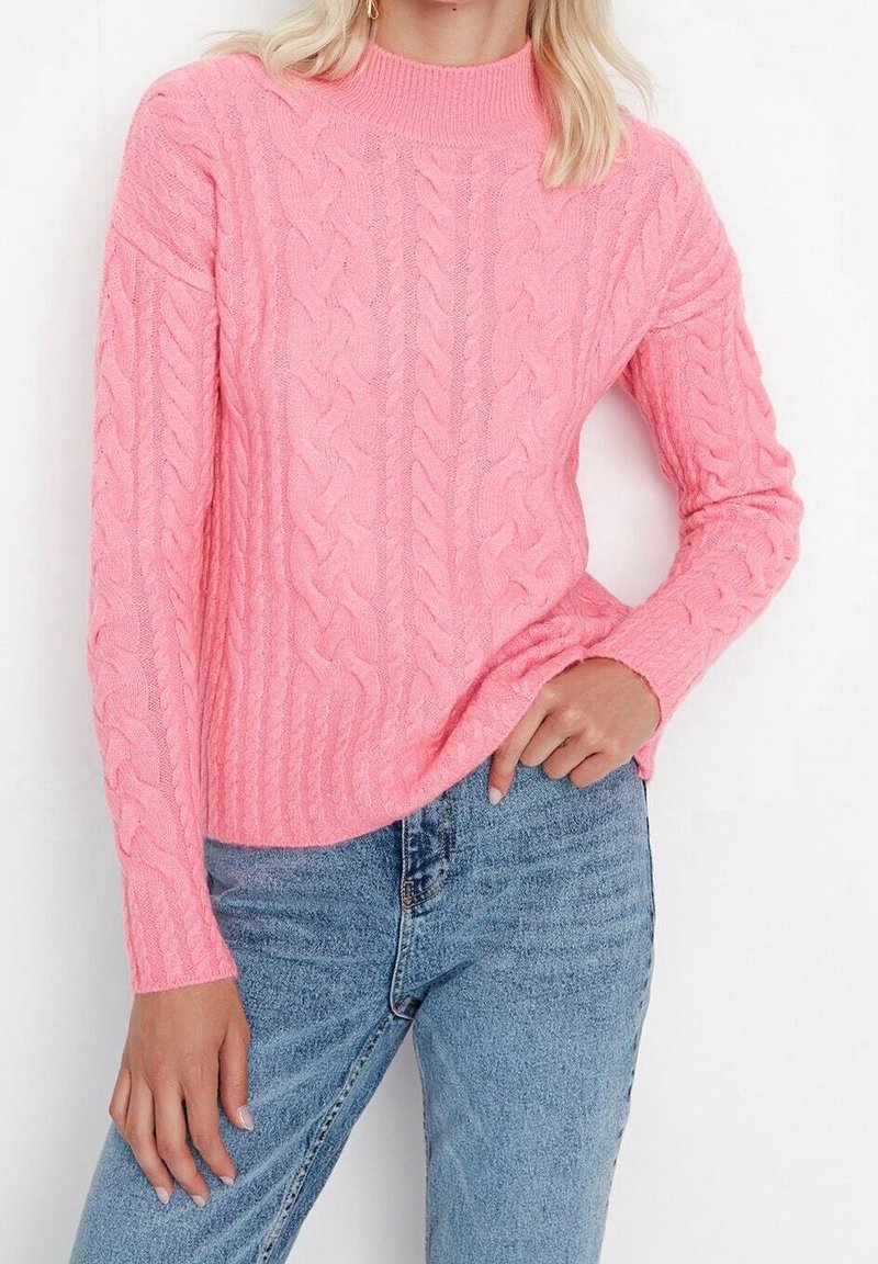Pull en tricot rose avec un col haut, présentant un motif texturé. Les manches sont longues et il est associé à un jean bleu.