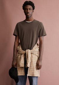 Braunes Baumwoll-T-Shirt, kariertes hellbraunes und blaues Tuch um die Taille gebunden, dunkle Jeans, schwarze Mütze in der Hand, vor einem pinken Hintergrund.