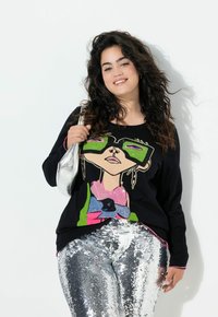 Czarny top z długim rękawem z kolorową grafiką twarzy postaci, zestawiony z błyszczącymi srebrnymi spodniami z cekinami i lśniącą srebrną torebką.