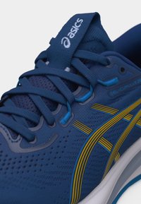 Κοντινή εικόνα μπλε παπουτσιού τρεξίματος ASICS GEL-CUMULUS 28 με κίτρινες ρίγες, κορδόνια και επάνω μέρος από αεριζόμενο δίχτυ.