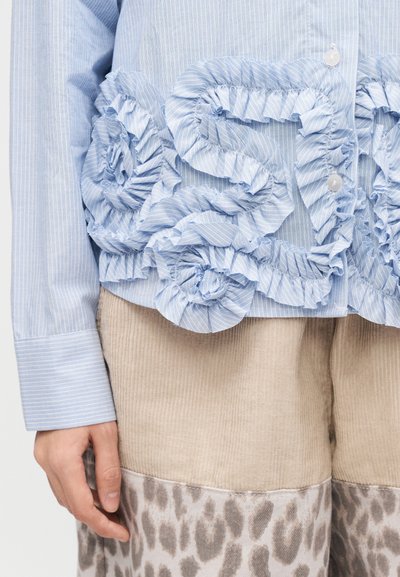 Ανοιχτό γαλάζιο πουκάμισο με ρίγες και μεγάλα, βολάν με λουλουδένιες λεπτομέρειες. Μακριά μανίκια με κλείσιμο με κουμπιά. Συνδυασμένο με μπεζ παντελόνι από κόντρα ύφασμα και διακοσμητικό ύφασμα με σχέδια.