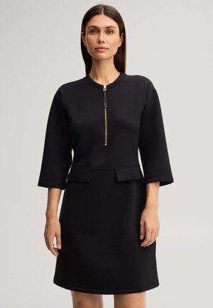 Donna con capelli scuri e lisci, che indossa un vestito nero con maniche tre quarti e una cerniera dorata dal collo al petto, in piedi contro uno sfondo semplice.