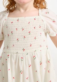 Hvid kjole med smocket bodice, korte puffærmer og spredte røde kirsebærmønstre. Den flydende nederdel tilføjer blødhed til designet.