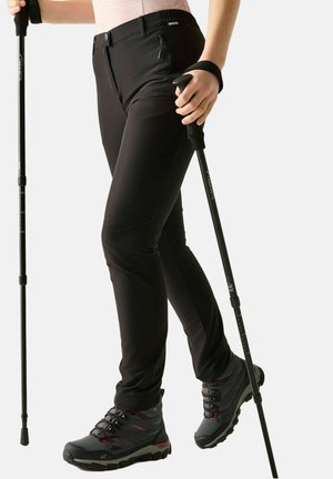 Persoon die zwarte broek en wandelschoenen draagt en zwarte verstelbare wandelstokken vasthoudt tegen een witte achtergrond.