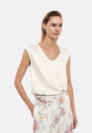 Donna con capelli chiari che indossa un top bianco senza maniche con scollo a V e pantaloni bianchi a vita alta con motivi floreali rosa e blu, in piedi con una mano in tasca.