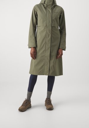 Long imperméable vert olive avec capuche porté sur des leggings bleu marine et des chaussures de randonnée marron avec des chaussettes grises épaisses.