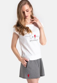 Biała koszula z krótkim rękawem z napisem „kochaj siebie” i czerwonym sercem, w połączeniu z czarno-białą spódnicą w kratę z czerwonym akcentem w kształcie serca.