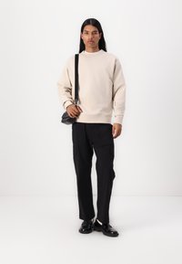 Sweatshirt bege com gola canelada, calças de carga pretas com bolsos e sapatos pretos. A pessoa transporta uma pequena mochila transversal preta.