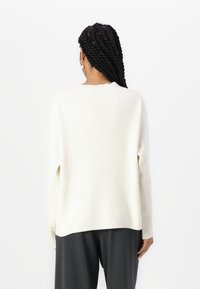 Pull en maille côtelée de couleur crème avec une coupe décontractée, un col rond et des épaules baissées, associé à un pantalon gris.