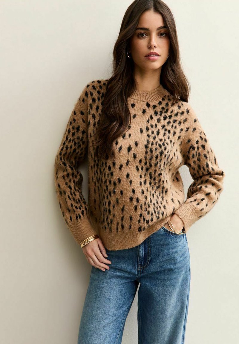 Suéter de punto beige con un patrón de manchas negras, mangas largas y cuello redondo. Combinado con jeans de mezclilla azules.