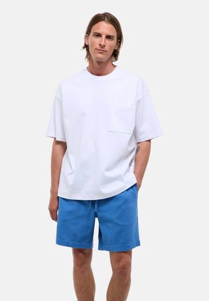 Jeune homme aux cheveux bruns mi-longs portant un t-shirt blanc oversize avec une poche et un short bleu décontracté, debout les mains détendues.